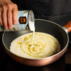 Person pouring a can of cream into a frying pan with a white substance, likely pancake batter.