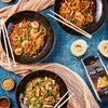 Three bowls of pad Thai with chopsticks on a blue tablecloth, featuring Shan brand packaging.